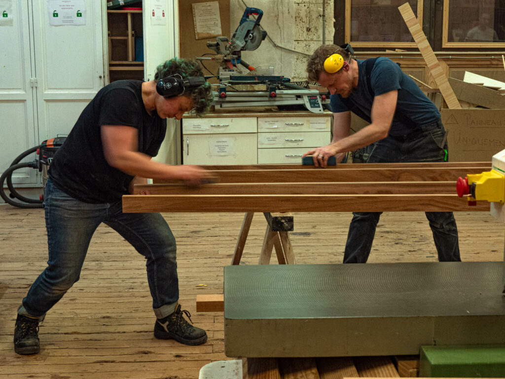 Deux apprenants en CAP Menuiserie à la Philomathique de Bordeaux concentrés sur le ponçage manuel d'une pièce en bois dans l'atelier.
